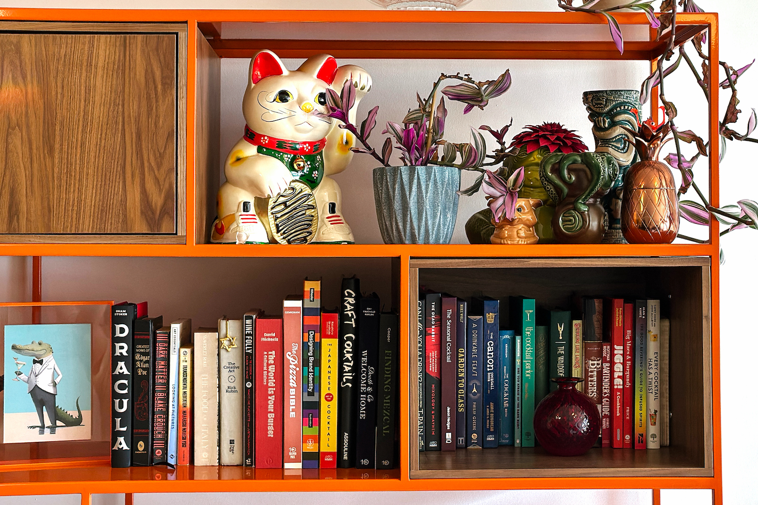 a bright orange and wooden bookshelf with books, plants, tiki mugs and a money cat